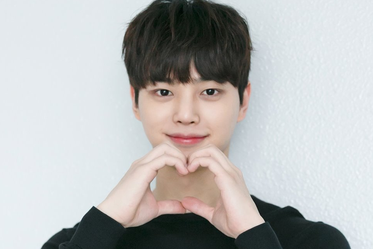 身穿黑色毛衣的男性，雙手做成心形放在胸前，白色牆壁為背景