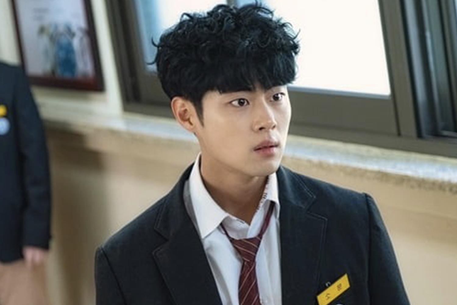穿著韓國高中制服的男性站在窗邊，襯衫配領帶，背景模糊顯示學校環境細節。