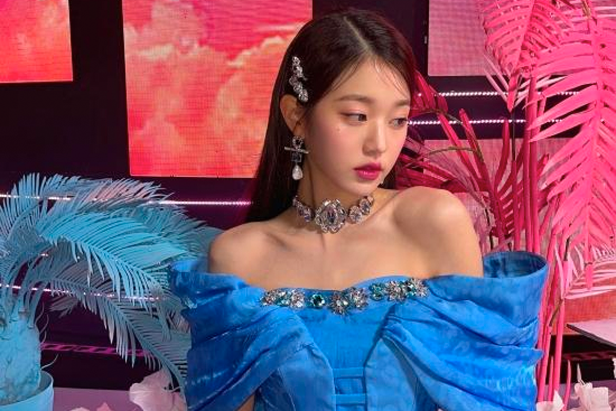 IVE成員員瑛身穿藍色華麗禮服，頸間戴著鑲嵌珠寶的項鍊，背景為粉紅色風扇葉裝飾。