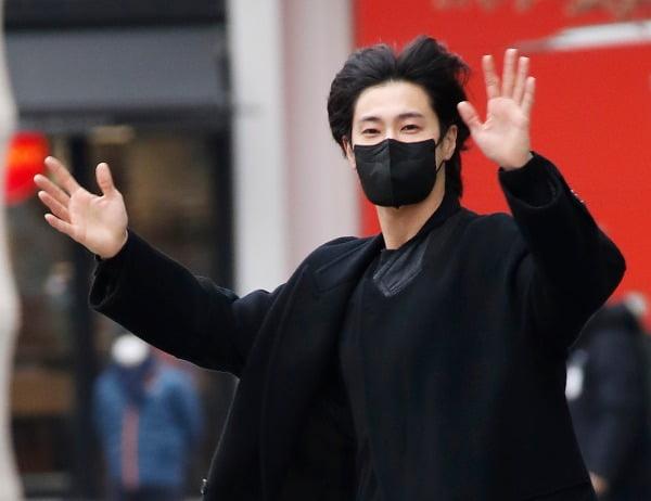 東方神起のメンバーが笑顔で手を振りながら、ソウルの放送局を出る姿。背景にはぼんやりと赤いカンバンが見える。冬の服装。