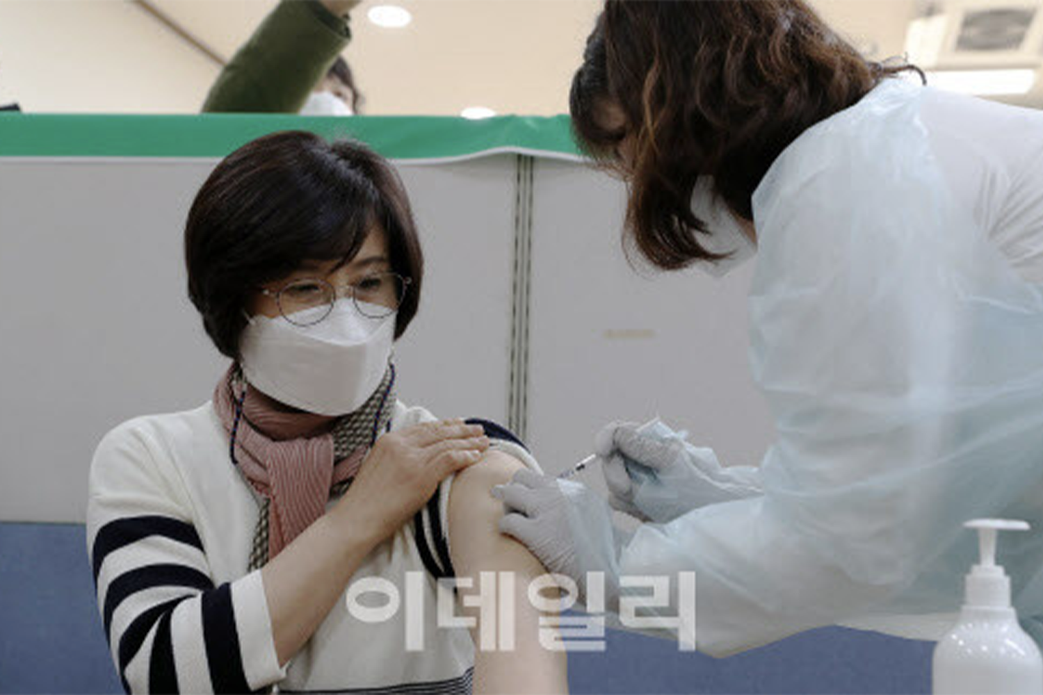 一名醫護人員正為一名坐著的女士接種新冠疫苗，女士身穿條紋毛衣，身旁有消毒液。出處：이데일리