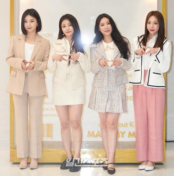 Brave Girlsが韓国のオンラインメディアイベントで、ビーズの背景に立ちハートの手を作っている、フォーマルな衣装を着たメンバーたち。