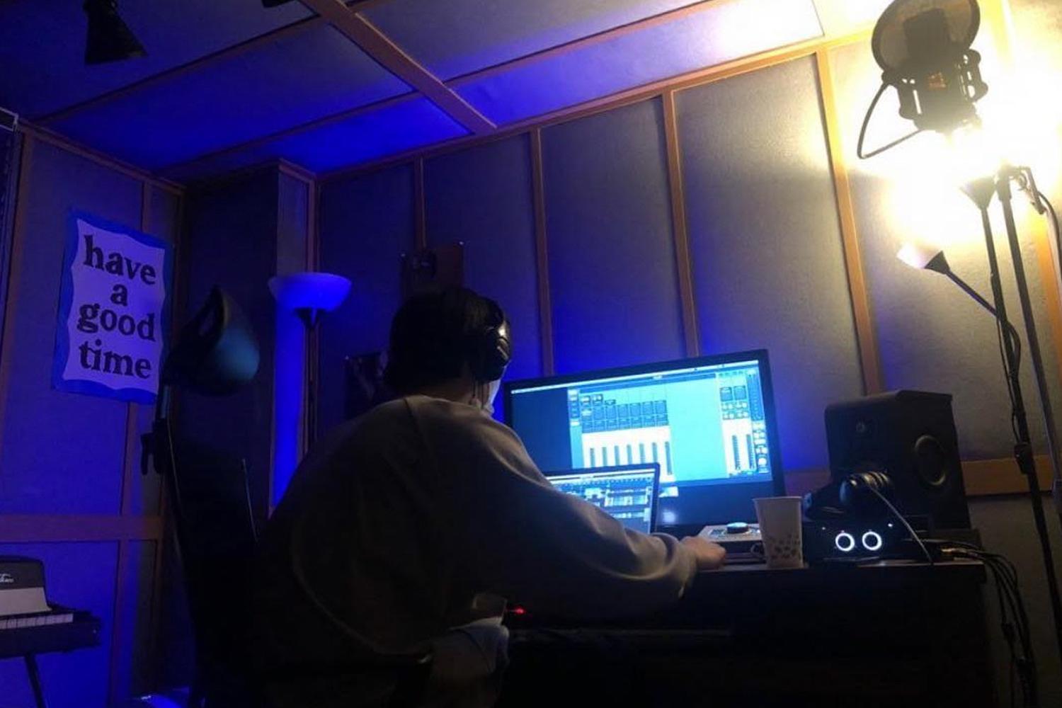 ชายหนุ่มนั่งทำงานอยู่ที่โต๊ะคอมพิวเตอร์ในสตูดิโอไฟสลัว หน้าจอคอมพิวเตอร์แสดงโปรแกรมควบคุมเสียง พร้อมอุปกรณ์ทำงานต่าง ๆ อาทิ หูฟังและไมโครโฟน