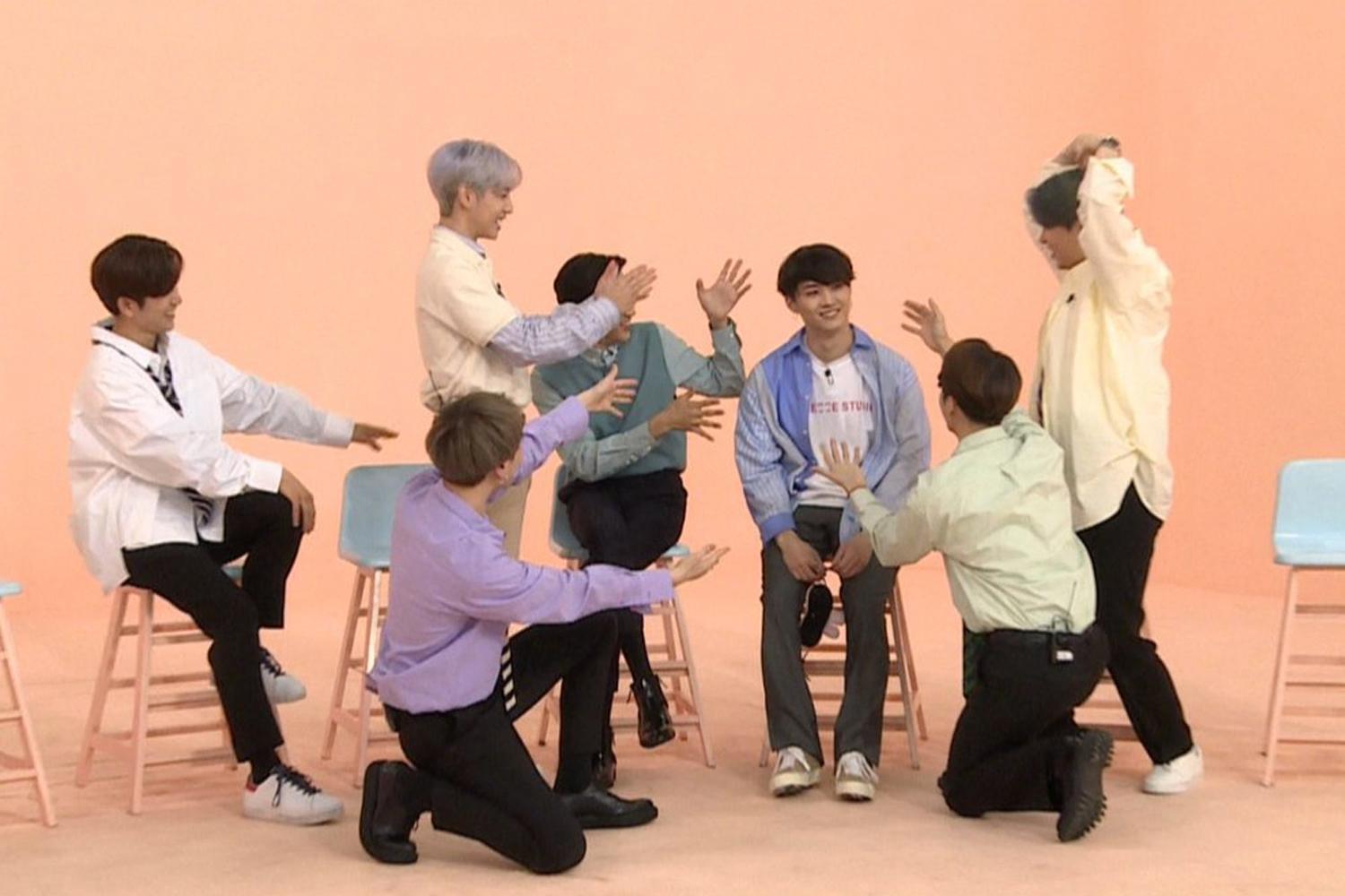 สมาชิก GOT7 แต่ละคนทำท่าทางที่หลากหลายในสตูดิโอสีชมพู เบื้องหน้าเป็นเก้าอี้สูงขนาดเล็ก บรรยากาศดูสนุกสนานและเป็นกันเอง