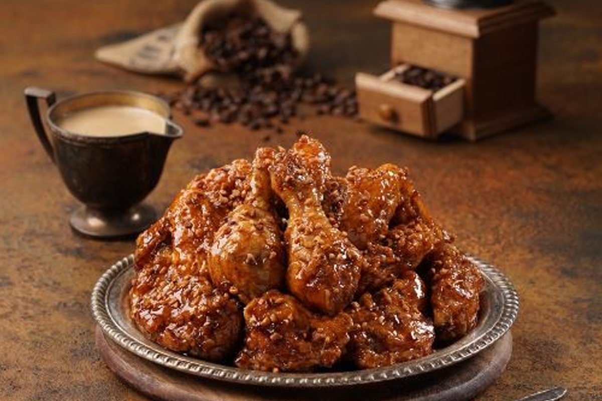 一盘装满咖啡拿铁风味炸鸡的银色盘子，旁边有一壶咖啡酱和咖啡豆，背景为原始木桌，衬托出传统与创新的彼此结合。