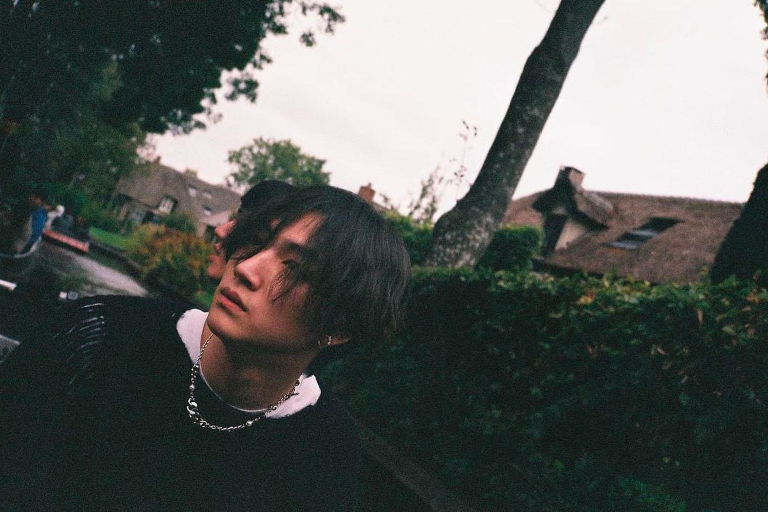 ศิลปินหนุ่มสวมสร้อยคอทองคำยืนอยู่ในสวนพร้อมเฟรมภาพท้องฟ้าหม่น