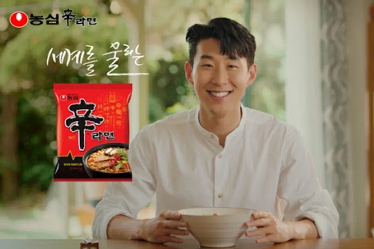 一名男子在桌前拿著泡麵碗，背景是辛拉麵產品包裝和品牌標誌。