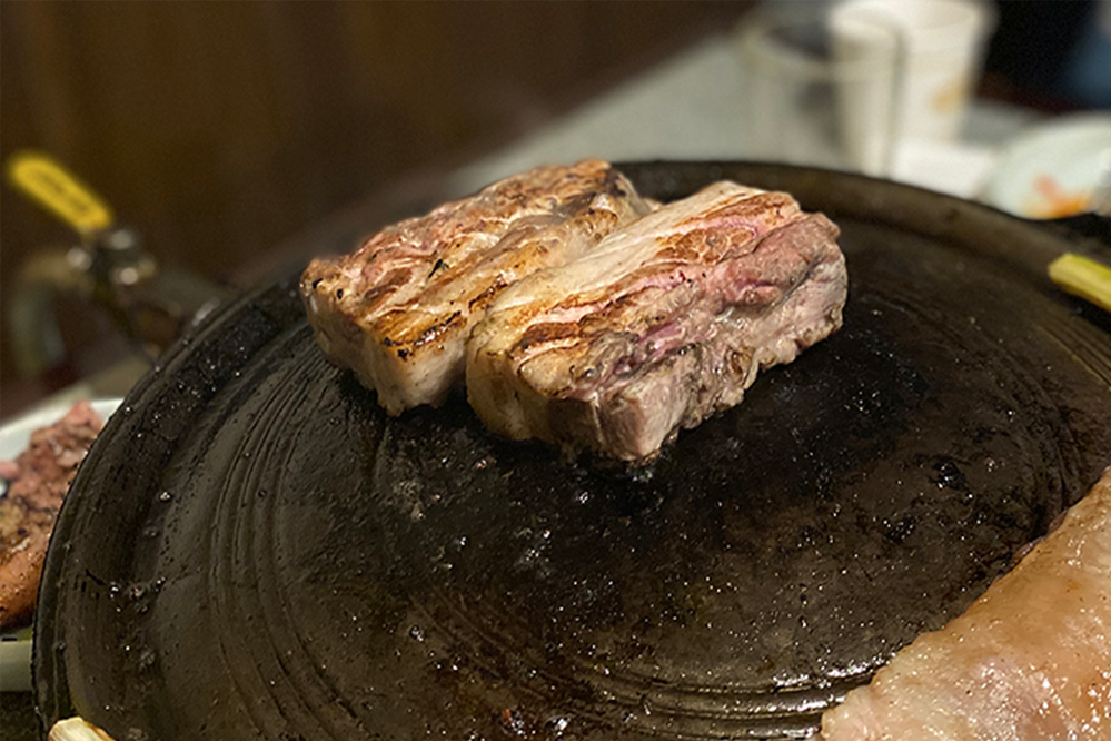 在梨泰院的夢炭餐廳，三層五花肉炭烤至完美金黃色澤，放在傳統鍋蓋中的特寫畫面，完美展示了韓式烤肉的美味特色。