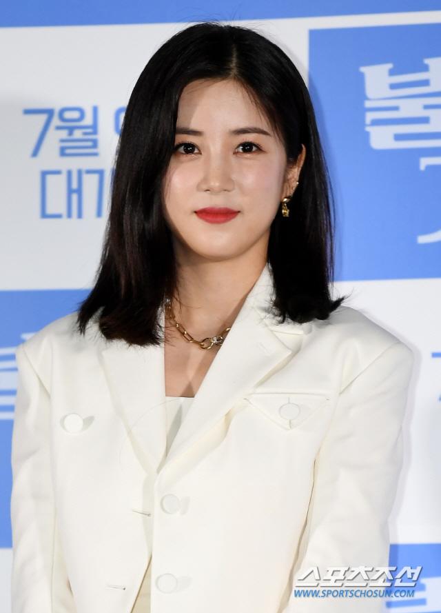 白いジャケットを着た女性が公の場に立っている。手にメモを持ち、淡々とした表情。青い背景には日付とイベント名が書かれている。