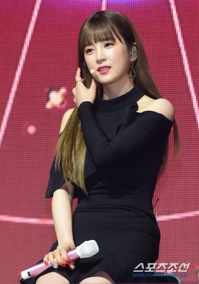 黒いドレスを着た女性がステージに座り、ピンク色のマイクを持っている。背景には大きな画面が映し出され、ステージ照明が雰囲気を作り出している。