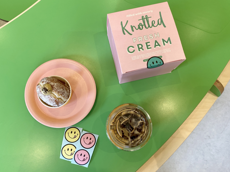 Розовая коробка Knotted с надписью 'СВЕЖИЕ СЛИВКИ', хол. американо и пончик на зелёном столе в кафе Knotted, Самсеонг, Сеул.