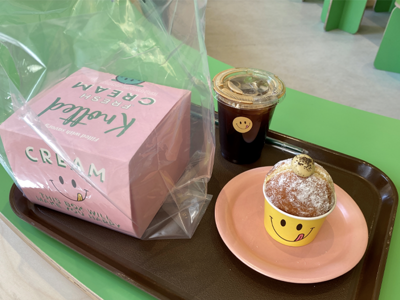 Набор из розовой коробочки с пончиками, чашки холодного американо и пончика в жёлтой упаковке с улыбающимся лицом на подносе в кафе Knotted, Сеул.