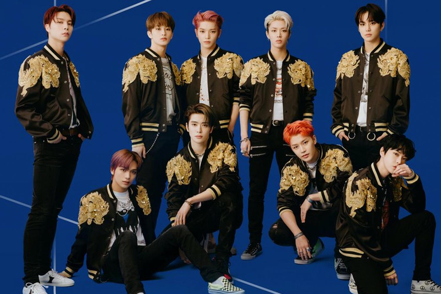 สมาชิกวง NCT 127 ในชุดแจ็กเก็ตสีดำและลวดลายทหารยืนล้อมหน้าแบ็คกราวน์สีน้ำเงิน