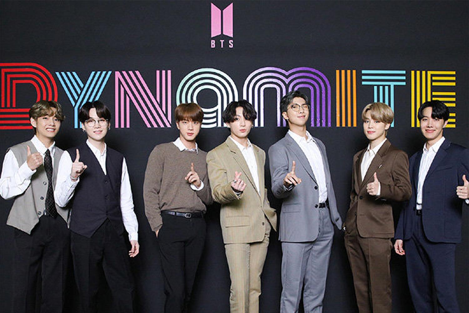 วง BTS สวมชุดสูทจัดแสดงในหน้าแบ็คกราวน์ที่มีกราฟิกตัวหนังสืออาร์ทของคำว่า 'DYNAMITE' สีรุ้ง