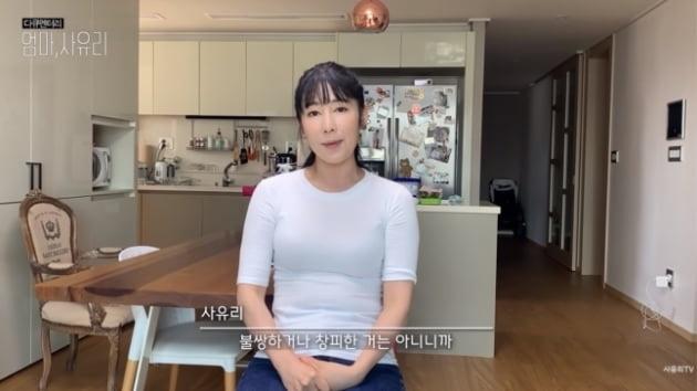 白いトップを着た女性、小百合のYouTube動画の一コマ。リビングルームのインテリアが背景にある。