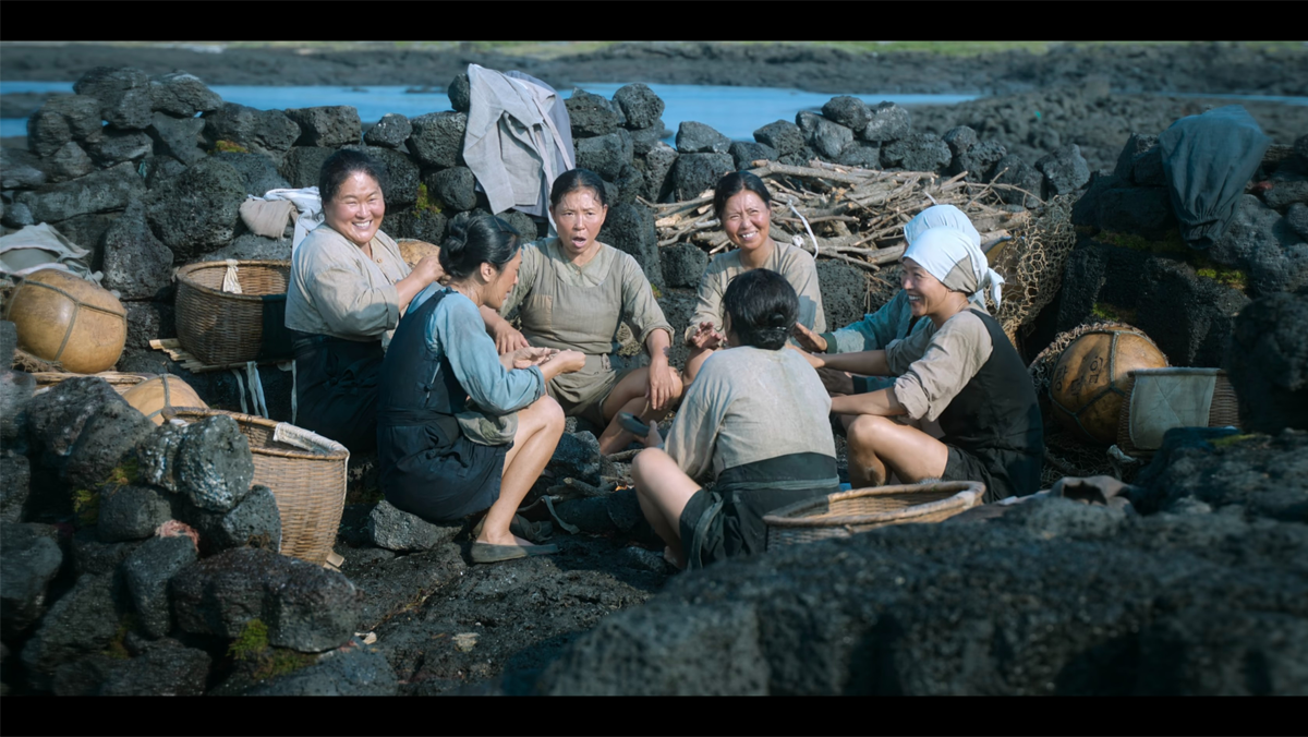 在石頭前坐著的濟州島海女群體圖像，展示海女的工作休息場景。