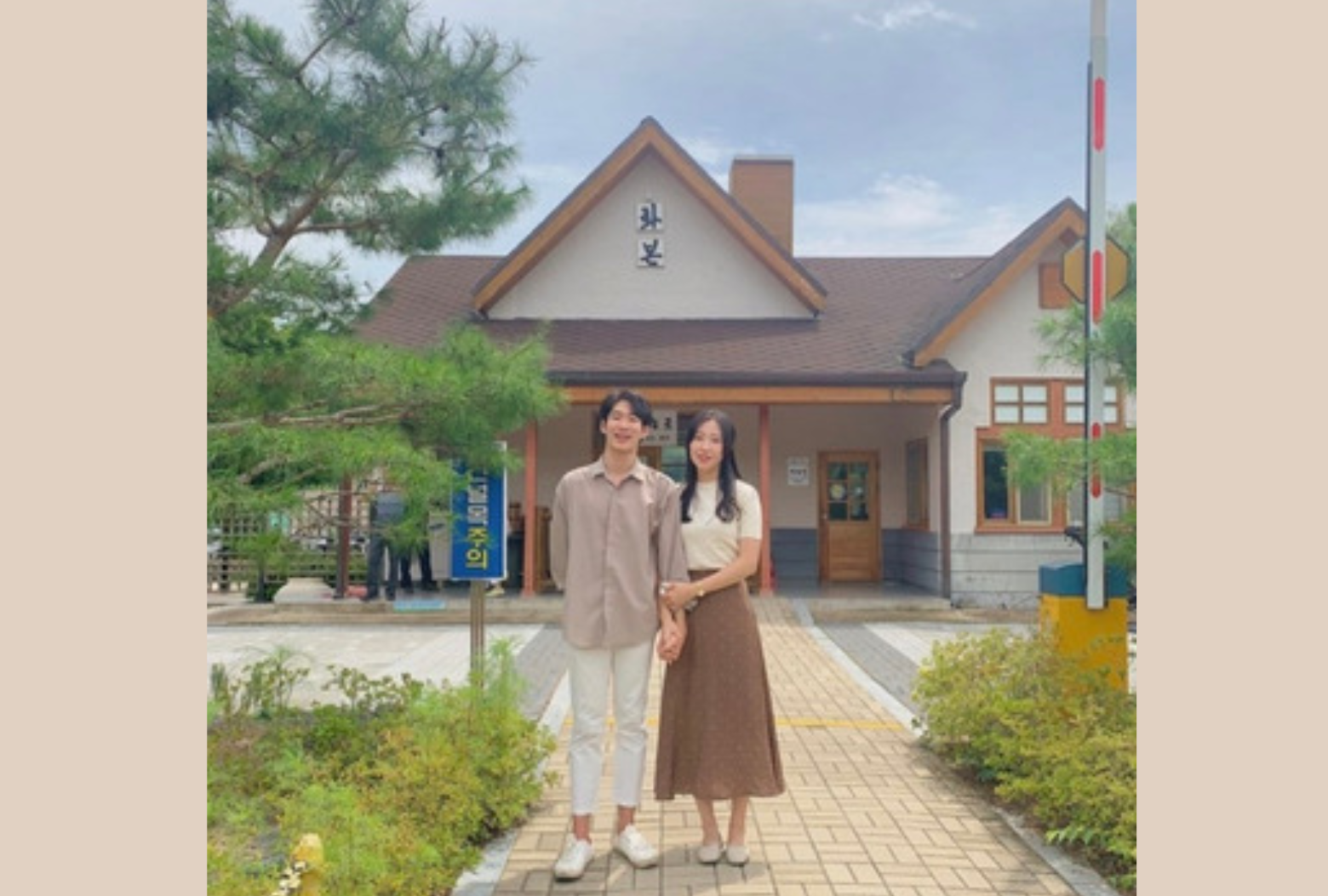 韓国軍威郡の花本駅の前景。駅の外観を前に、二人が手を繋いでポーズを取っている。緑豊かな駅の周辺が見える。