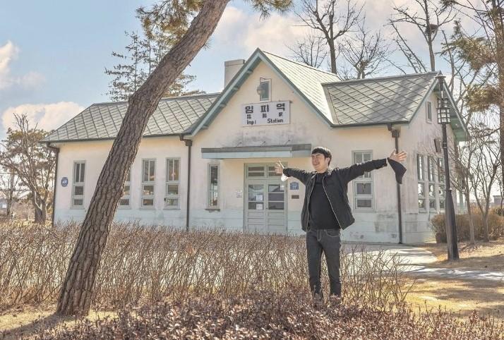 韓国群山市の臨陂駅を背景にした人物が両手を広げているシーン。水色の壁と特徴的な屋根の駅が見える。