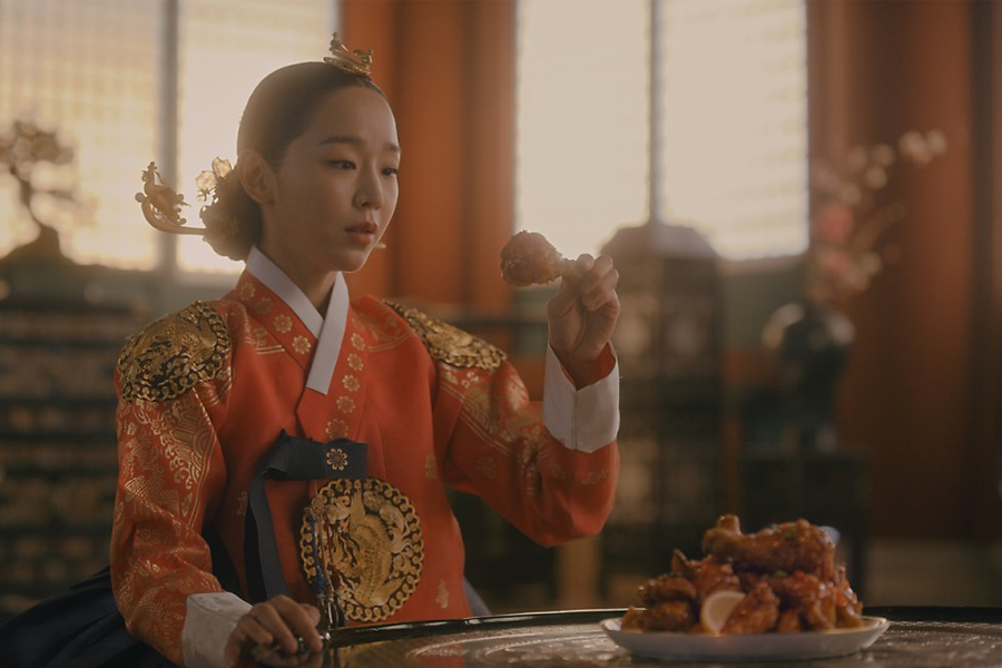 穿着传统韩服的代言人坐在餐桌前吃炸鸡，场景设计为古代宫殿风格，背景是暖色的室内装饰。