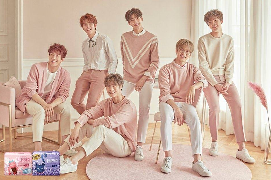 韩国男子团体在卫生棉广告中穿着粉色和白色服装，坐在客厅环境中，产品展示在画面左下方。
