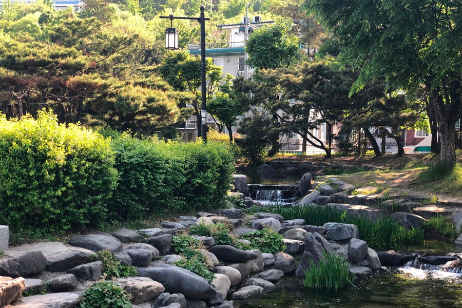 A scenic stream flows through Namsan Hanok Village, surrounded by bushes, trees, and rocks for a tranquil setting.