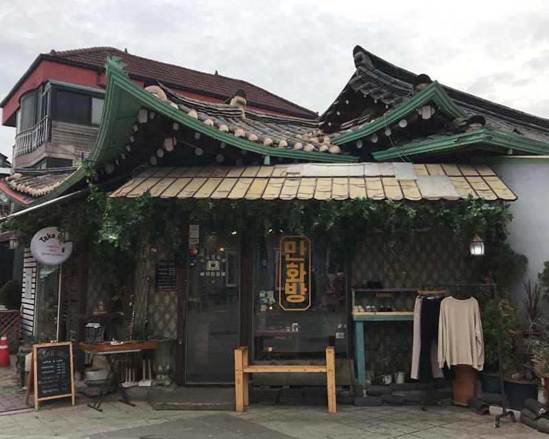 韓国の伝統的な屋根のあるカフェの外観。植物で飾られた入り口の横には看板があり、壁の一部には洋服が掛けられています。市街地の中に位置しています。