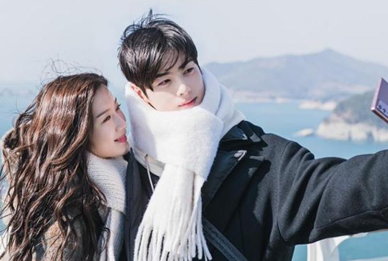 Zwei Menschen stehen eng beieinander, beide in Winterschals eingehüllt, während sie eine malerische Landschaft mit einer Küste im Hintergrund betrachten.