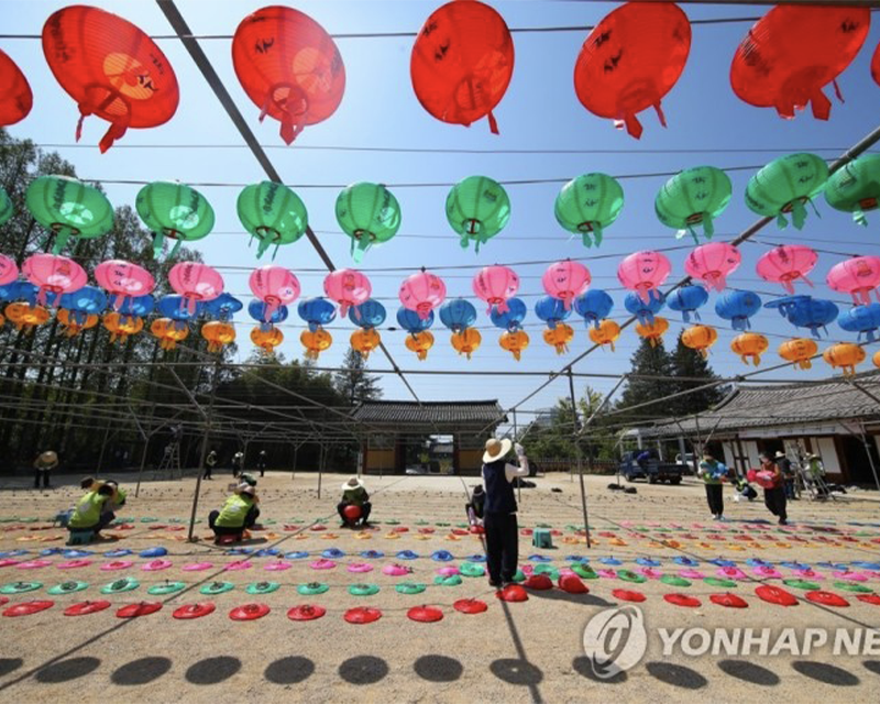 首爾奉恩寺一景色，藍天下懸掛著多彩的傳統佛教花燈，地面上工作人員正在佈置燈飾；這個場景象徵著佛誕節期間的慶祝準備和社區協作的畫面。