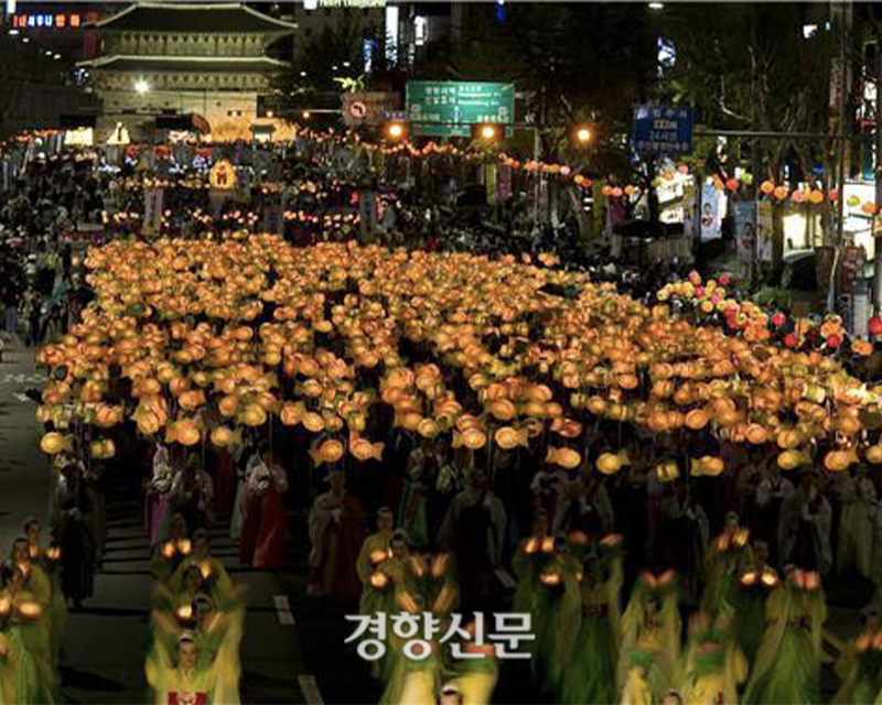 首爾市中心夜間佛誕節花燈遊行，參加者手持橙色花燈沿路行進，背景為城市街道和擠滿燈籠的頭頂，展現炫麗的燈光景觀，慶祝佛誕的歡樂氣氛。