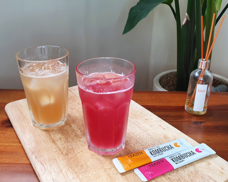 Una imagen de dos vasos de kombucha TEAZEN en la mesa, uno de sabor limón y otro de bayas, junto a sus respectivas barras de embalaje en un tablero de madera.