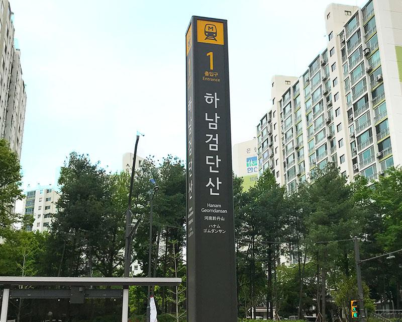 韓國京畿道河南黔丹山地鐵站1號出口的標誌，背景是高樓和樹木。
