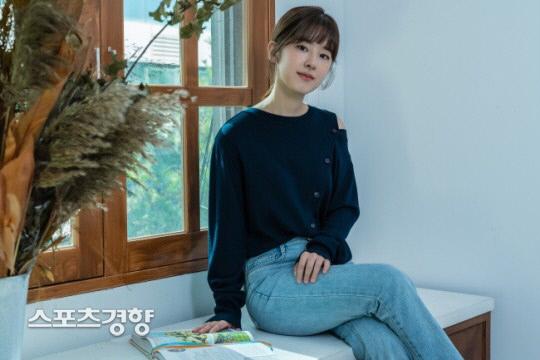 青いセーターを着てジーンズを履き、窓辺の白いベンチに座る女性の姿。背景には緑豊かな外の風景が見える。手元には開かれている書籍が置かれている。