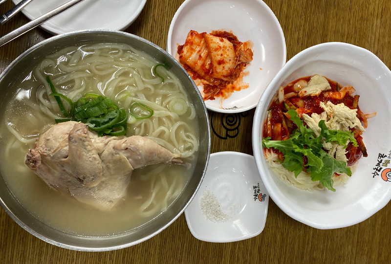 미사리 초계국수 본점에서 제공하는 초계비빔국수와 닭칼국수 요리입니다. 국수와 닭 고기가 담긴 국물 및 매운 소스를 얹은 면 요리가 식탁에 정렬되어 있습니다. 생김치와 소금, 간장도 포함된 한상의 모습입니다.