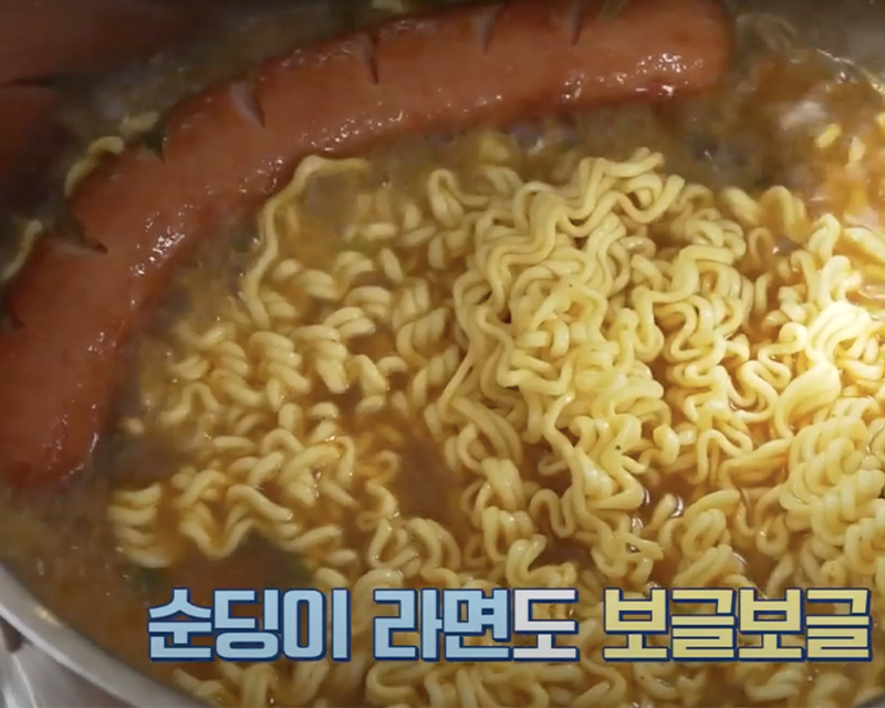 Mie ramyeon yang sedang mendidih di dalam panci, disajikan dengan sosis dan kuah ramen. Gambar menunjukkan sudut mendekat dari mie kuning keemasan, menonjolkan kelezatannya.