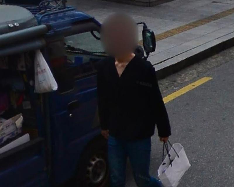 街道旁的藍色小卡車停泊在商業區，一名攜帶紙袋的路人沿著道路緩緩行走，背景是現代城市建築物的外觀。