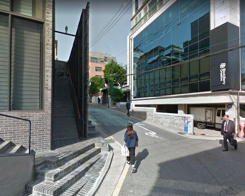 BlockBerry Creative 建築物旁的坡道，長髮女士背對著鏡頭走向公司方向，背景是商業建築和樹木。