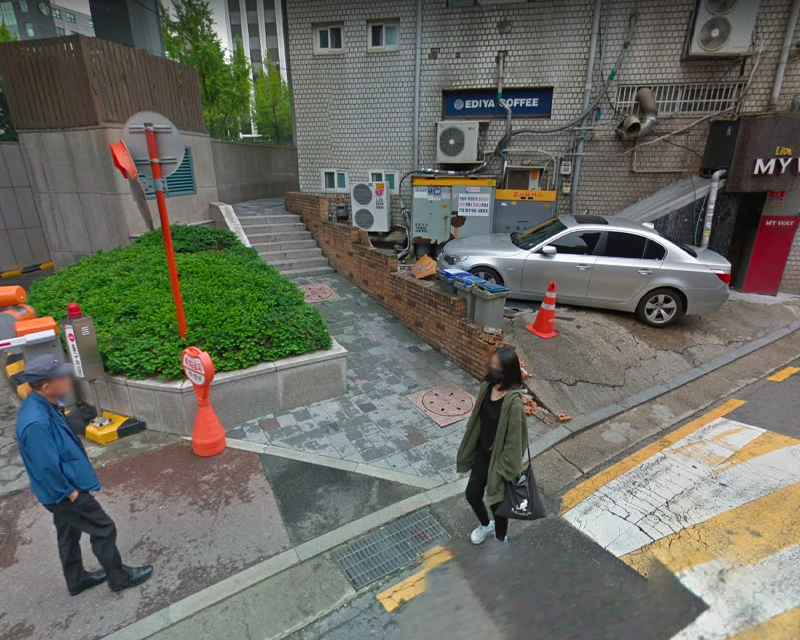 JYP 公司附近，銀色車輛停在樹籬旁，兩名路人，穿著深色服裝的女士攜帶黑色手袋，綠色植物裝飾環境。