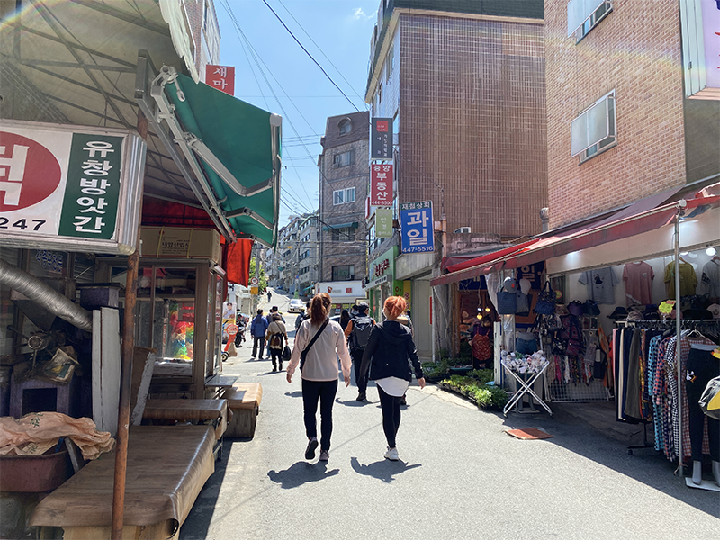 韓國首爾某時尚小巷的街景，服裝店陳列著五顏六色的商品，吸引行人駐足。