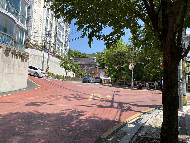 韓國首爾某小巷，行人在樹蔭下漫步，街道周圍的店家與招牌展現出當地的市井氣息。