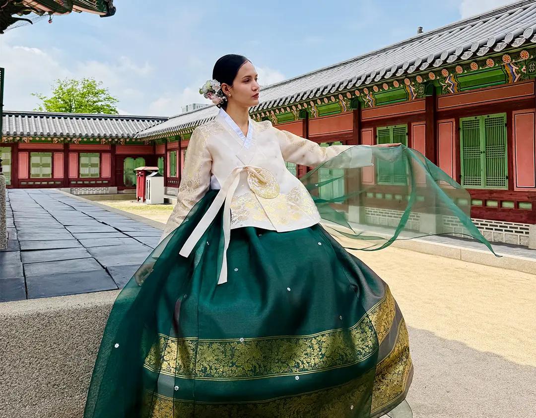 女性身穿綠色與金色刺繡韓服站在景福宮院落，薄紗隨風飄起，傳統宮牆與瓦片背景呈現首爾古宮白天典雅氛圍