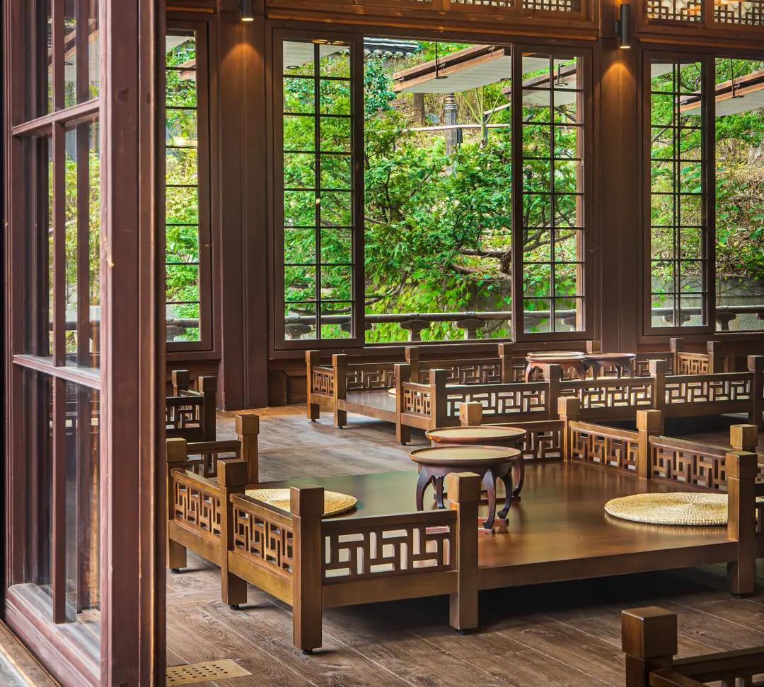 景福宮附近傳統茶館的室內一隅，木質地板與雕花矮桌、坐墊排列整齊，大窗外綠意盎然，適合穿韓服夜間參觀前的休憩拍照場景。
