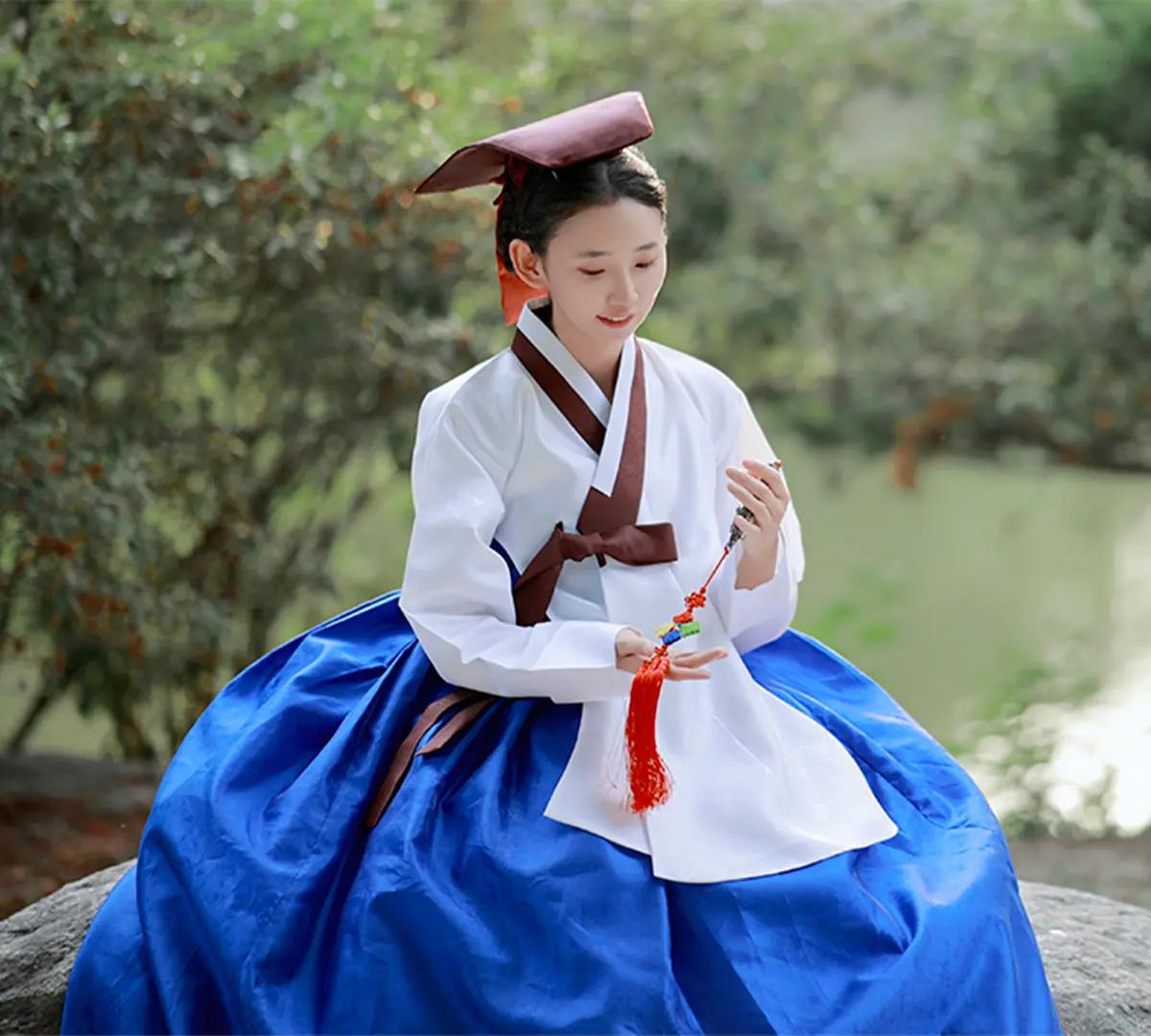 身著白色上衣與藍色裙裝的女性，佩戴傳統韓國帽飾與手持流蘇飾品，坐在庭院石上，在景福宮周邊的綠意池畔拍照，展現韓服夜間入場氛圍