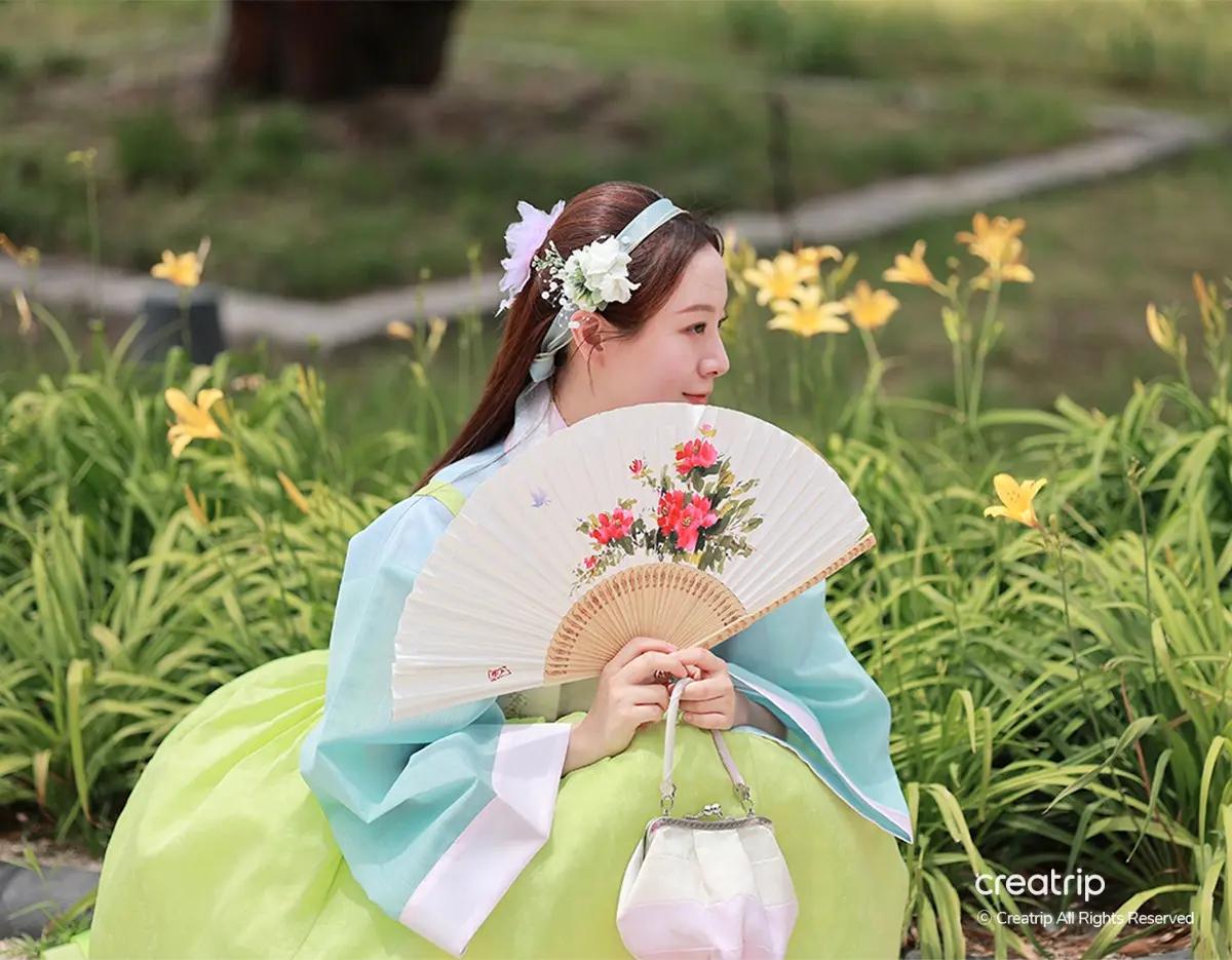 身穿淺藍綠韓服的遊客坐在花叢前，手持繪花折扇與小手提包，背景為景福宮夜間開放附近的庭園與黃花，突顯韓服拍照與夜間參觀情境