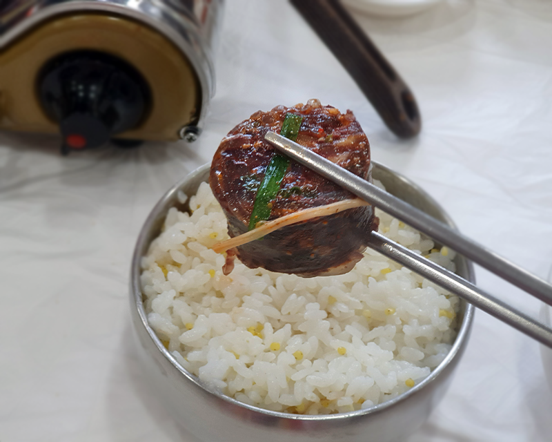 Holding garlic sundae with silver chopsticks above a bowl of rice at Chungcheongdo Sundae