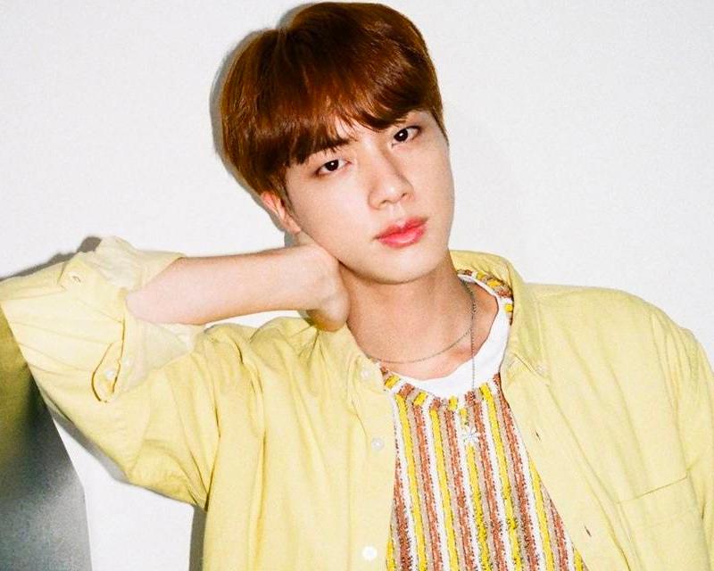 穿著白色長袖的Jin坐在椅子上，靠著沙發，背景是簡單的房間設計，展現輕鬆儀態。