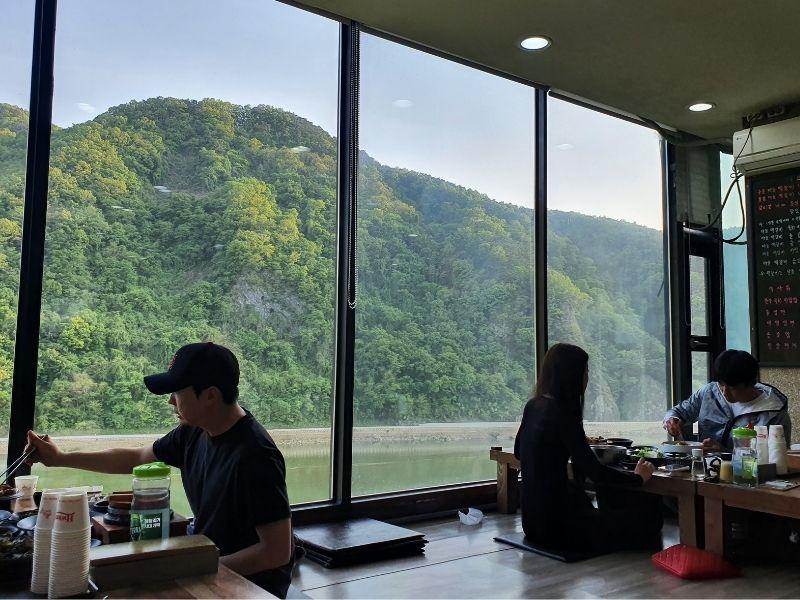 People grilling their food sitting on the floor next to a large window at Dawon Restaurant Danyang
