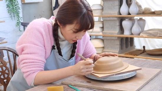 一位穿著粉色毛衣的人正在陶藝工作室裡製作陶盆,桌上有海綿和工具,環境整潔舒適。