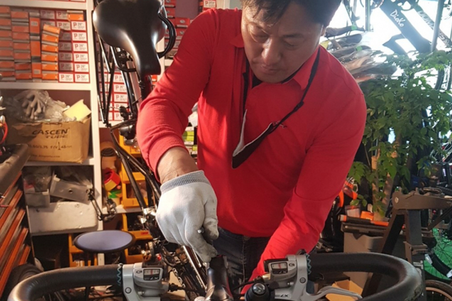 穿著紅色上衣的人正在修理自行車,在工作間裡使用工具專注工作,周圍擺滿了修理相關設備。