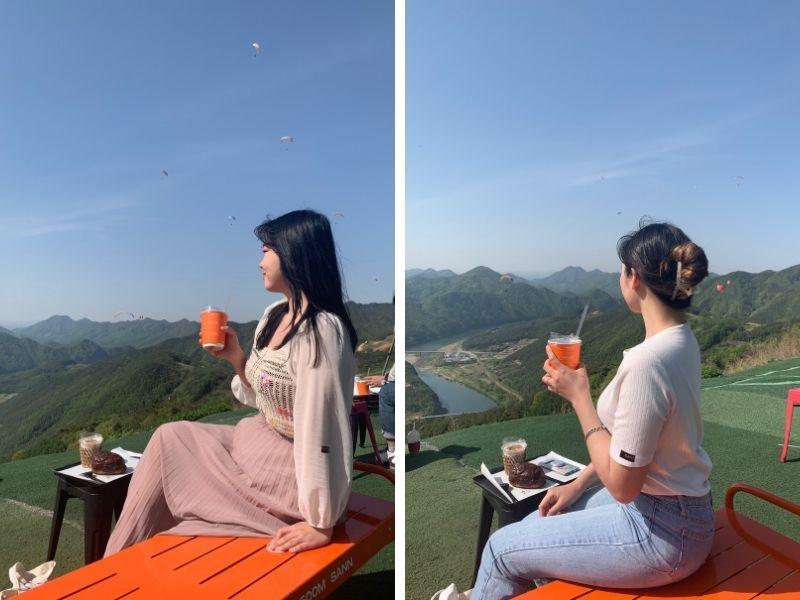 side by side images creatrip editors posing with orange togo cups of coffee in hand sitting on orange bench with paragliders and mountains in background in danyang south korea