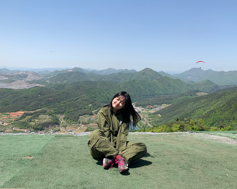 creatrip editor sitting on mountain ledge with blue sky, mountains, town, and paragliders in the background in danyang south korea